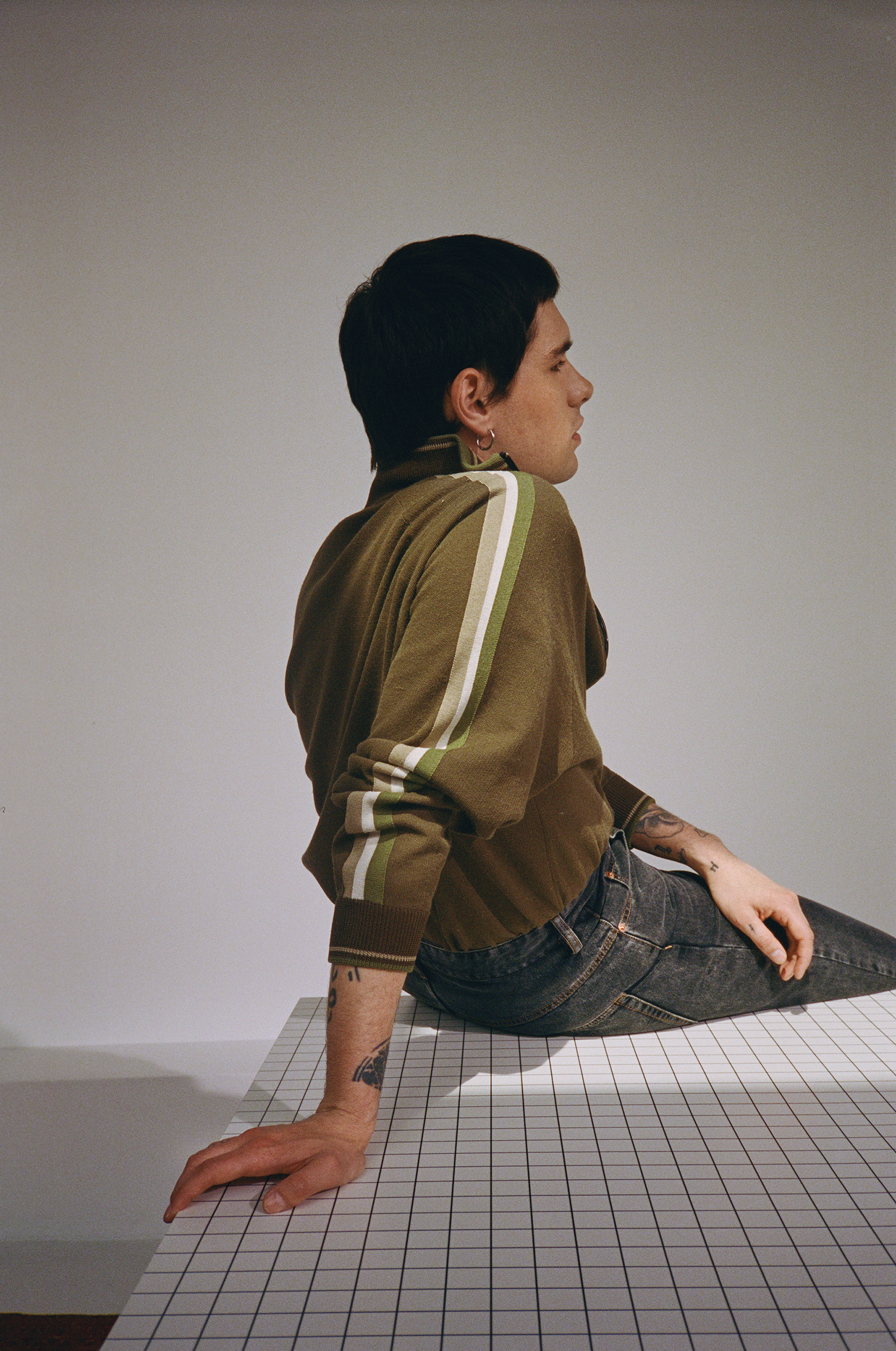 Profile of a young adult male with dark hair wearing an olive green long sleeve shirt and jeans, sitting on a white checkered surface.