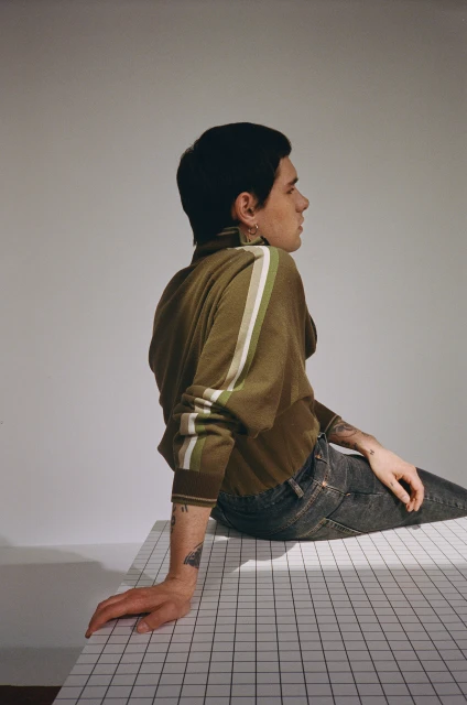 Profile of a young adult male with dark hair wearing an olive green long sleeve shirt and jeans, sitting on a white checkered surface.