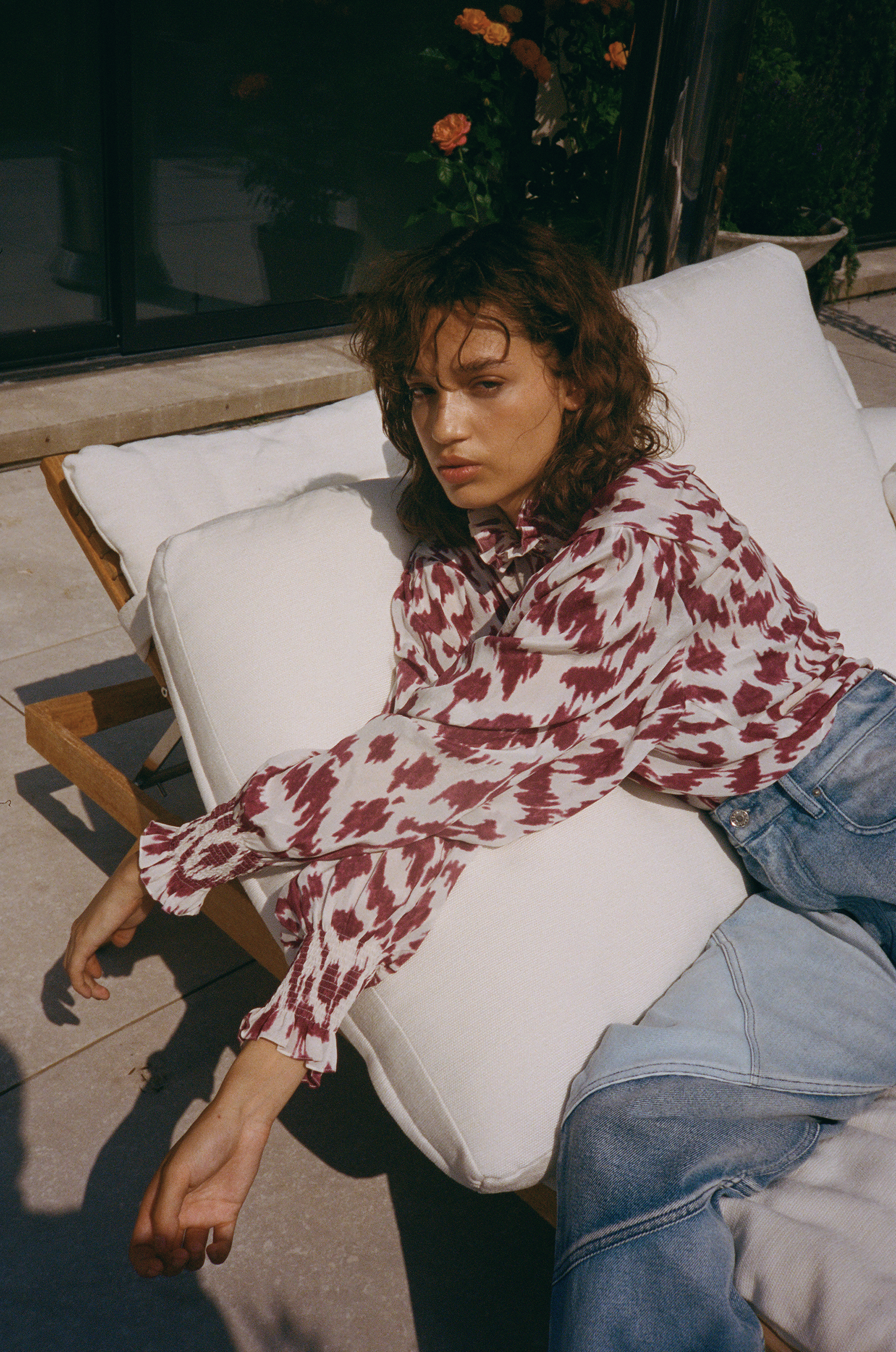 A woman with curly brown hair reclining on a white outdoor lounge chair wearing a patterned blouse and denim jeans.