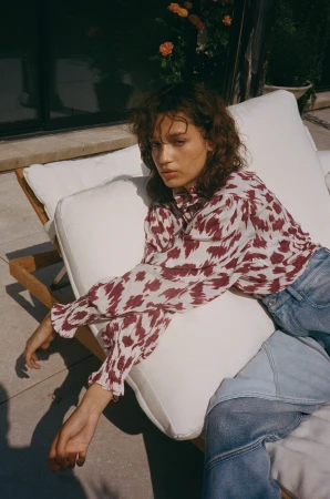 A woman with curly brown hair reclining on a white outdoor lounge chair wearing a patterned blouse and denim jeans.