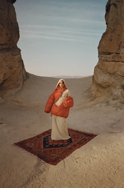 Woman wearing a large orange puffer jacket and head covering, standing on a red patterned rug in a desert landscape between rock formations.
