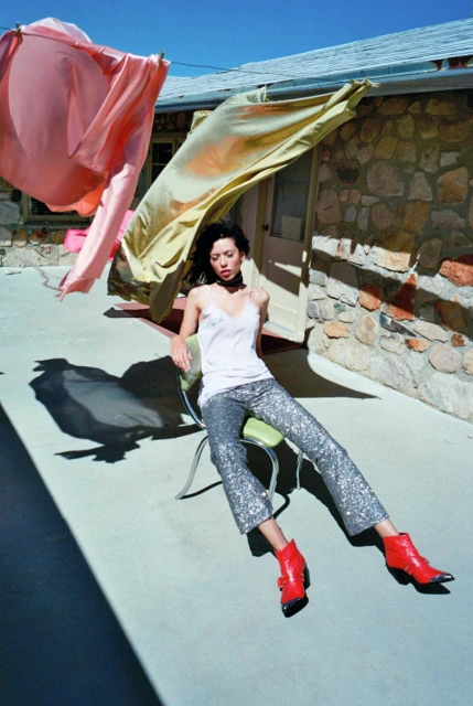 Fashion shot: A woman in a white top and sequin pants sits on a chair outside on a concrete patio, with fabric hanging from a clothesline.