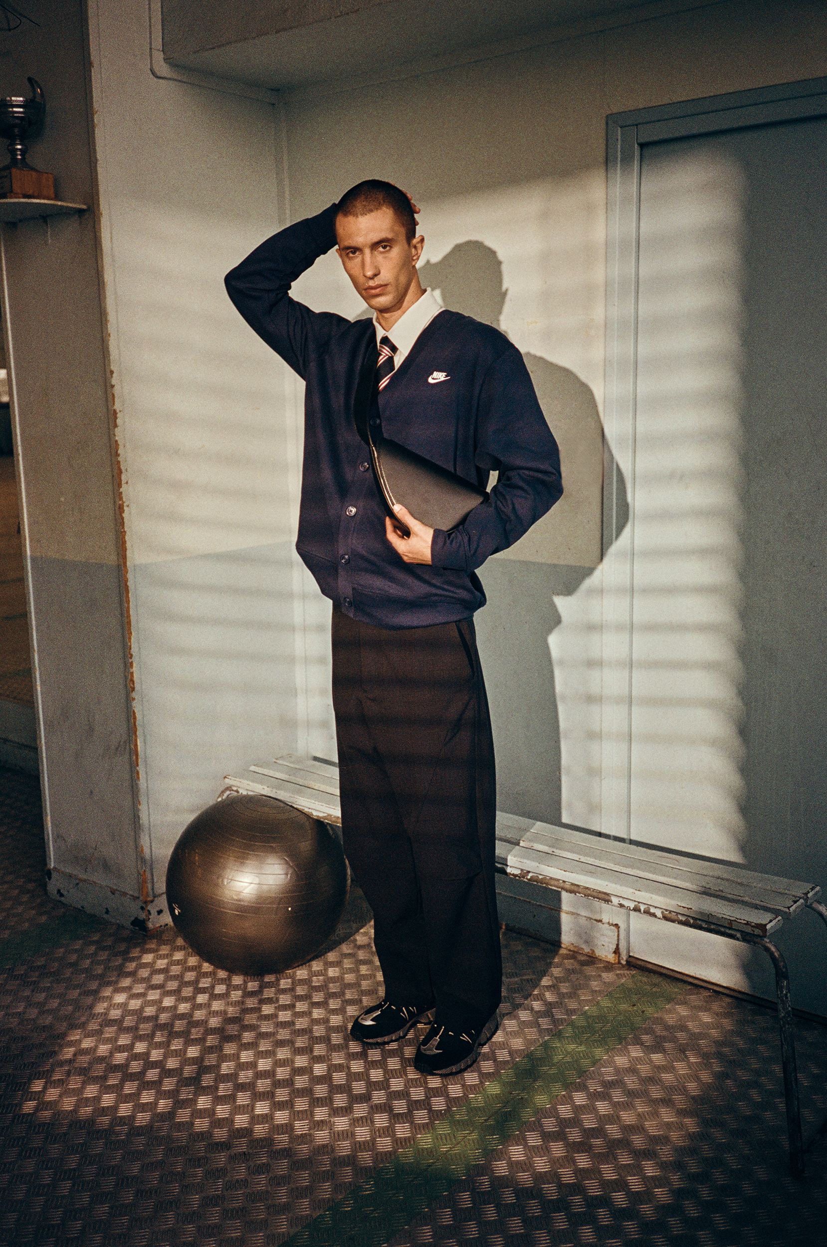 Man in cardigan and dress pants with athletic ball and bench in locker room-like indoor setting with slatted window shadows.