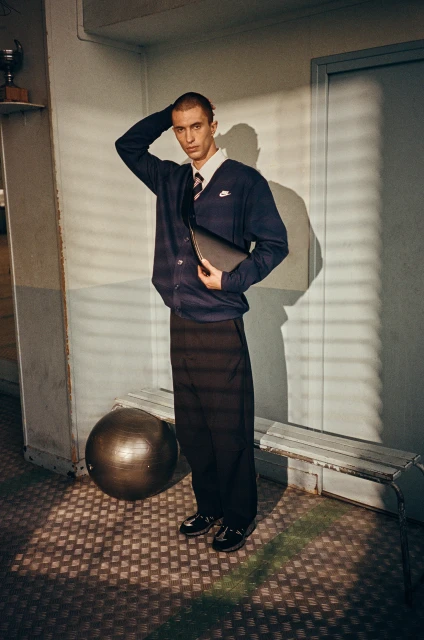 Man in cardigan and dress pants with athletic ball and bench in locker room-like indoor setting with slatted window shadows.
