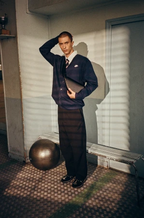 Man in cardigan and dress pants with athletic ball and bench in locker room-like indoor setting with slatted window shadows.