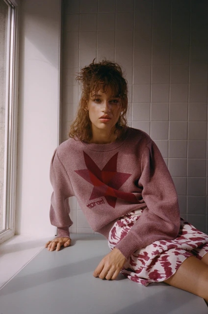 A female model in a mauve sweater with a star graphic and a patterned skirt poses near a window with natural light indoors.