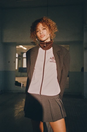 Woman with curly blonde hair wearing a grey blazer, white zip-up jacket, and grey pleated miniskirt.