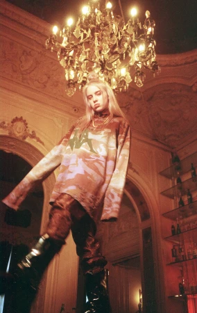 Low angle shot of Billie Eilish standing under an ornate crystal chandelier wearing a patterned long sleeve shirt and dark trousers.