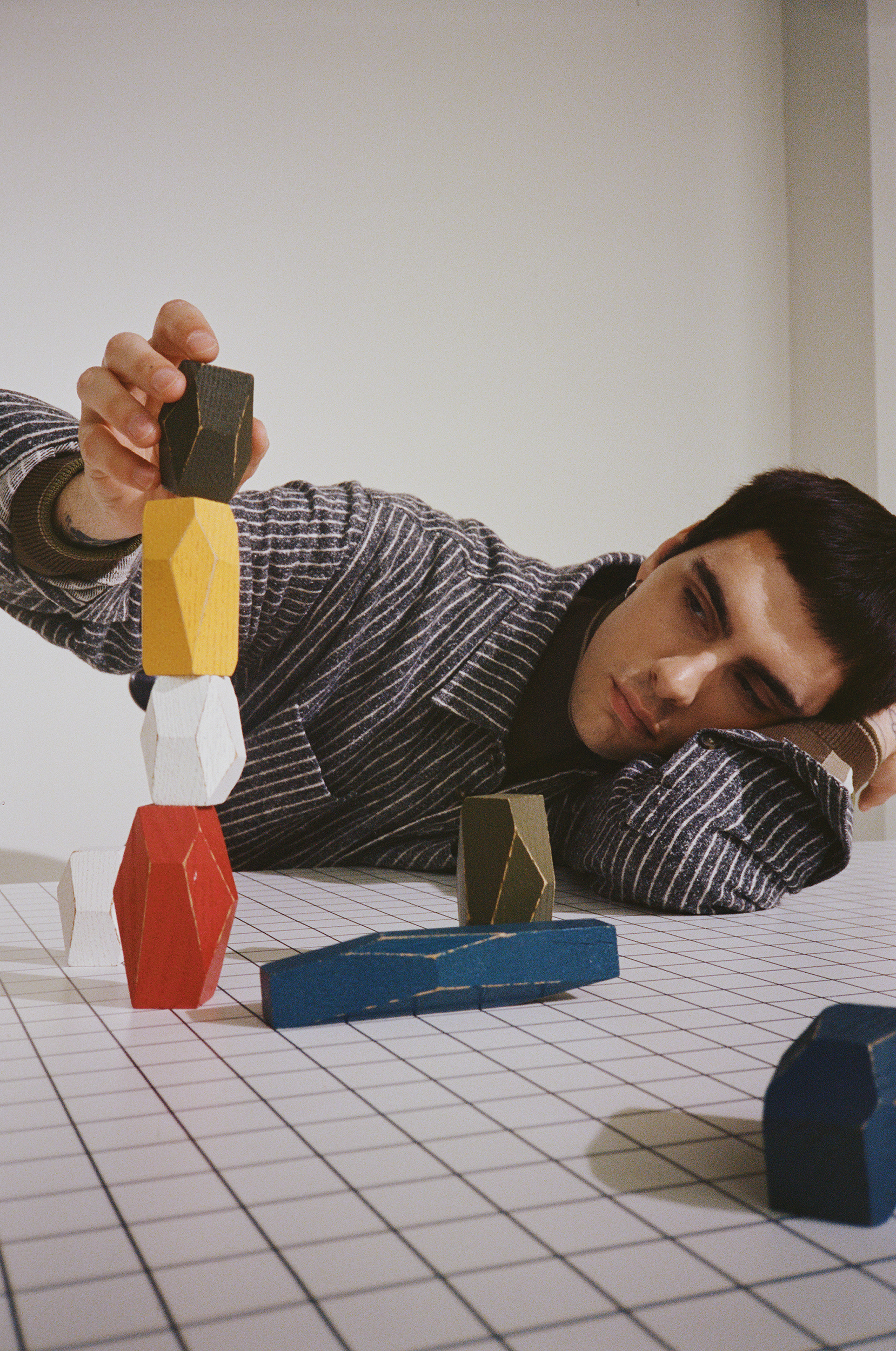 Man stacks colorful wooden blocks on a grid surface in a bright indoor space, wearing a striped shirt and dark undershirt with a serious expression.