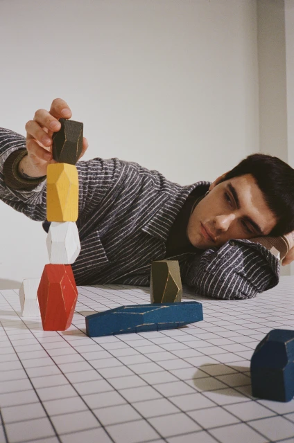 Man stacks colorful wooden blocks on a grid surface in a bright indoor space, wearing a striped shirt and dark undershirt with a serious expression.