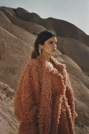 Woman wearing a textured rust-colored coat and elaborate ear cuff standing in front of barren, rolling sand hills