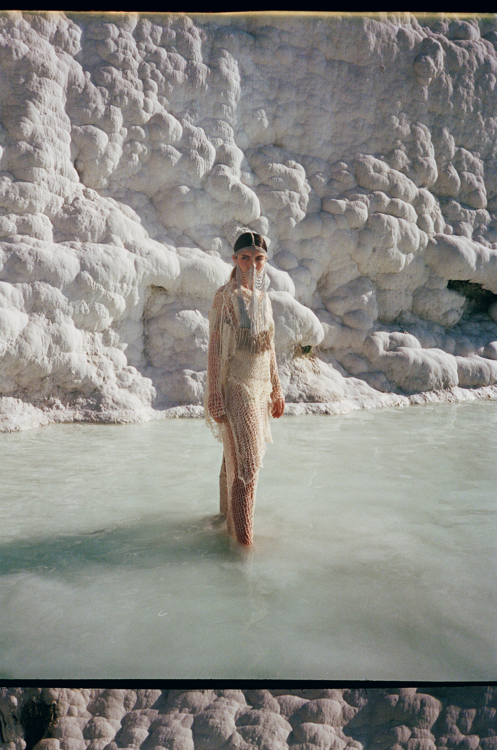 Adult female in a light dress stands in shallow turquoise water surrounded by textured white travertine formations on a sunny day.