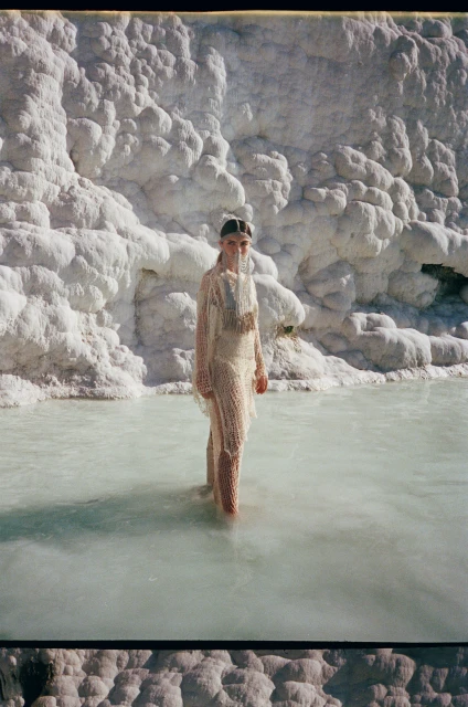 Adult female in a light dress stands in shallow turquoise water surrounded by textured white travertine formations on a sunny day.