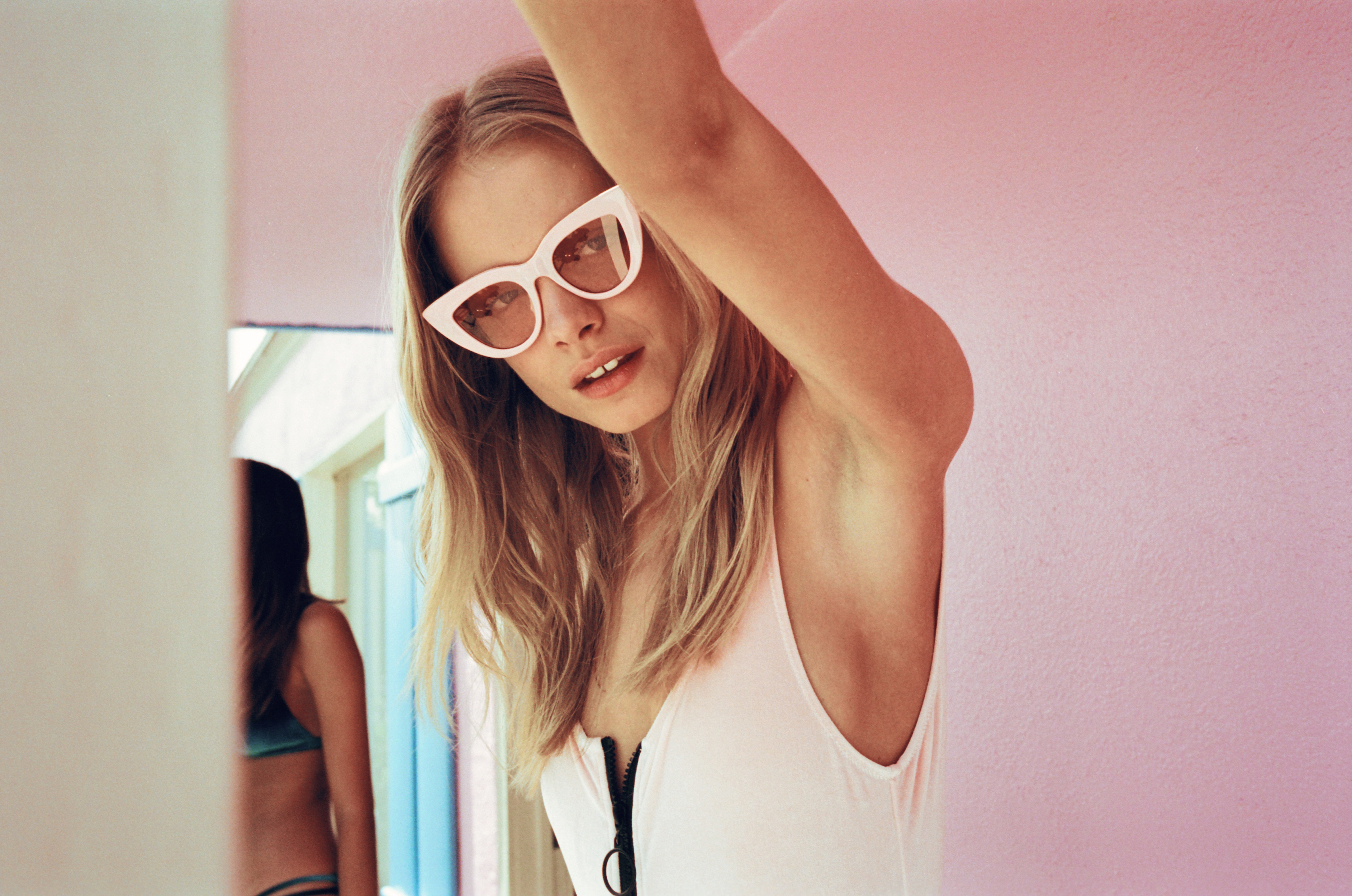 Close-up of a blonde woman with white sunglasses wearing a white one-piece swimsuit, posing indoors with a pastel-colored background.
