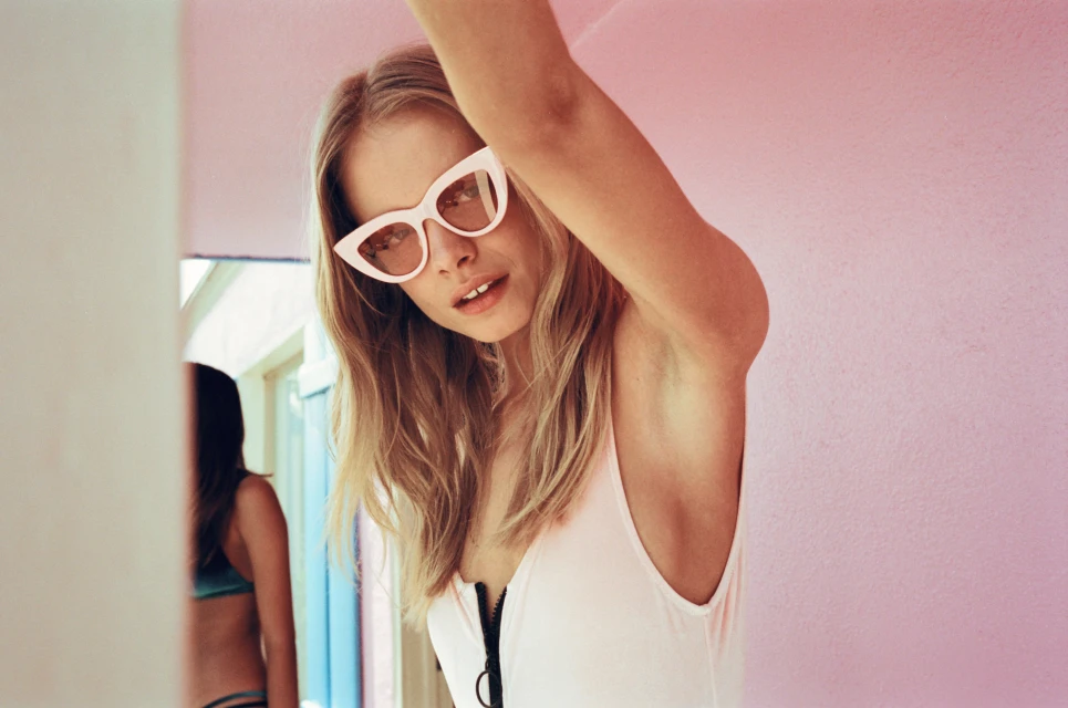 Close-up of a blonde woman with white sunglasses wearing a white one-piece swimsuit, posing indoors with a pastel-colored background.