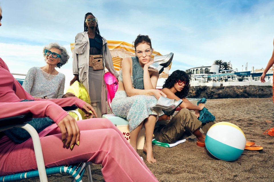 A group of women in fashionable clothing are gathered on a beach on a sunny day, with a building visible in the background.
