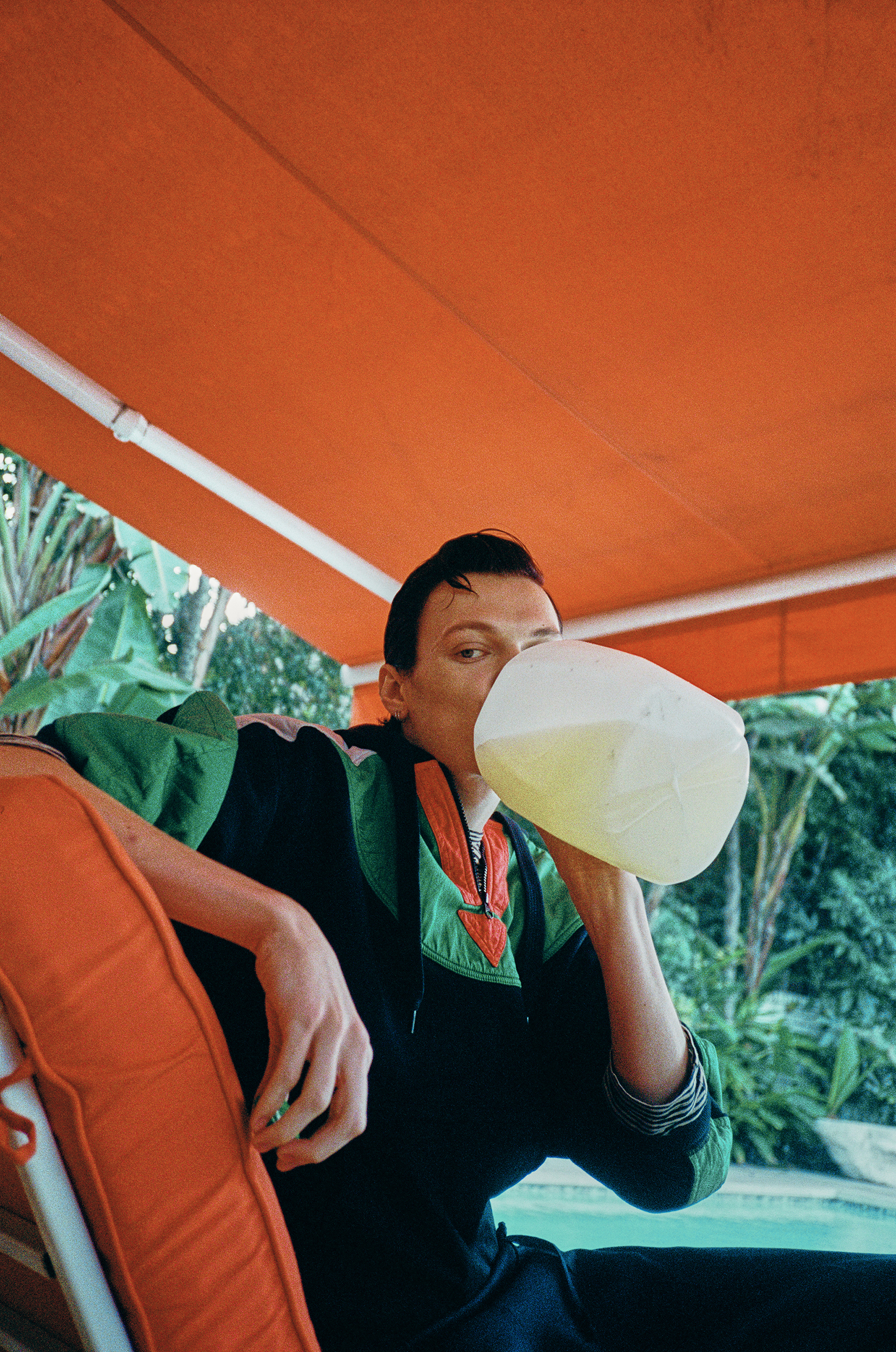 A person drinks from a large jug outdoors. The individual is seated on a patio chair under an orange awning, surrounded by lush greenery.