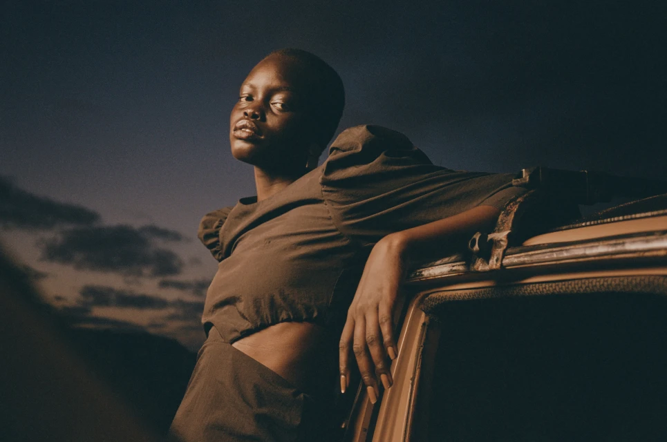 Stylish portrait of a Black woman in a brown dress leaning against a vintage car under a dramatic blue and cloudy sky at twilight.