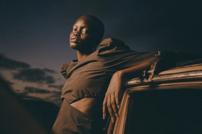 Stylish portrait of a Black woman in a brown dress leaning against a vintage car under a dramatic blue and cloudy sky at twilight.