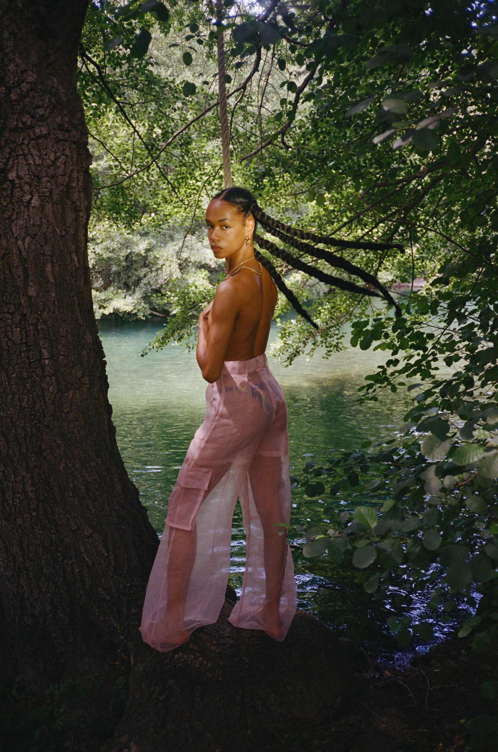 Woman poses barefoot on a tree trunk in a forest, wearing sheer light pink pants with her back exposed, looking over her shoulder.