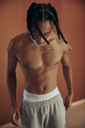 Man with bare torso and braided hair looks down in a medium shot against an orange wall wearing grey sweatpants and a pearl necklace.