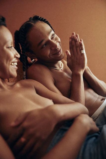 Intimate portrait of a smiling couple with braids, jewelry, and bare chests, lying closely in front of a plain terracotta backdrop.