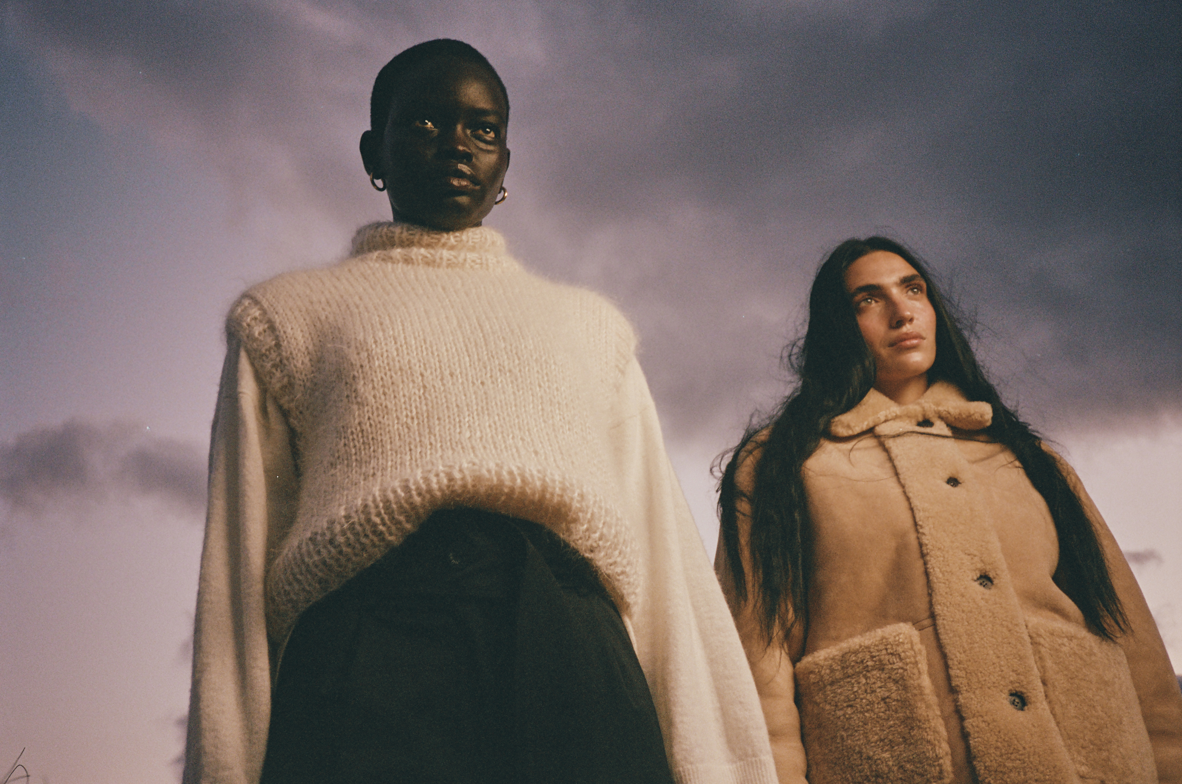 Two models wearing knitwear and shearling coats, standing outdoors with a cloudy sky backdrop. Fashion editorial shot with natural lighting.