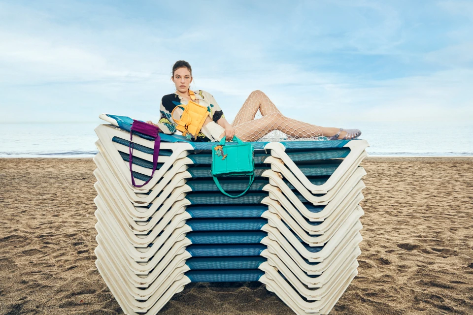 An adult female model lounges on a stack of white and blue beach chairs on a tan sandy beach with pastel blue skies and ocean backdrop.