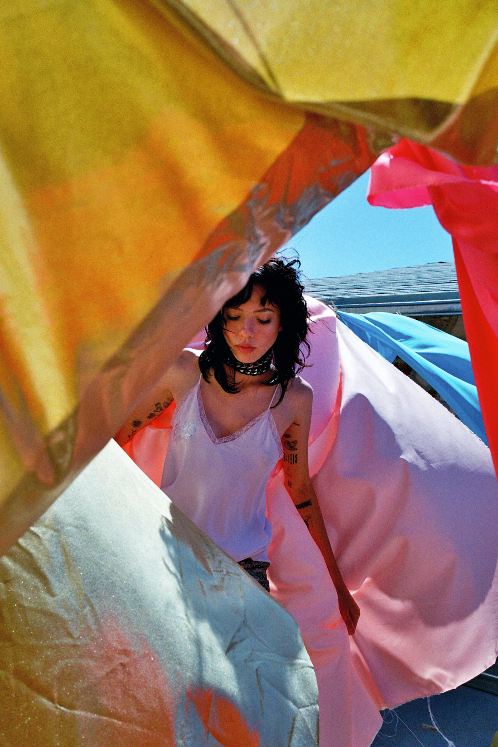 A young adult woman with short dark hair stands outside under a colorful array of fabrics in the daylight with a choker necklace.