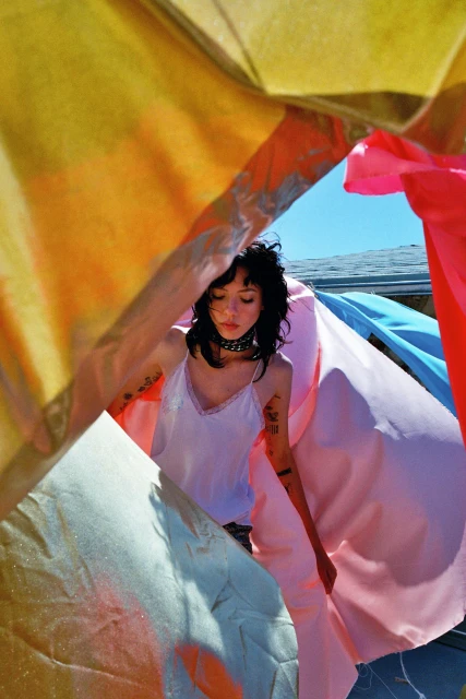 A young adult woman with short dark hair stands outside under a colorful array of fabrics in the daylight with a choker necklace.
