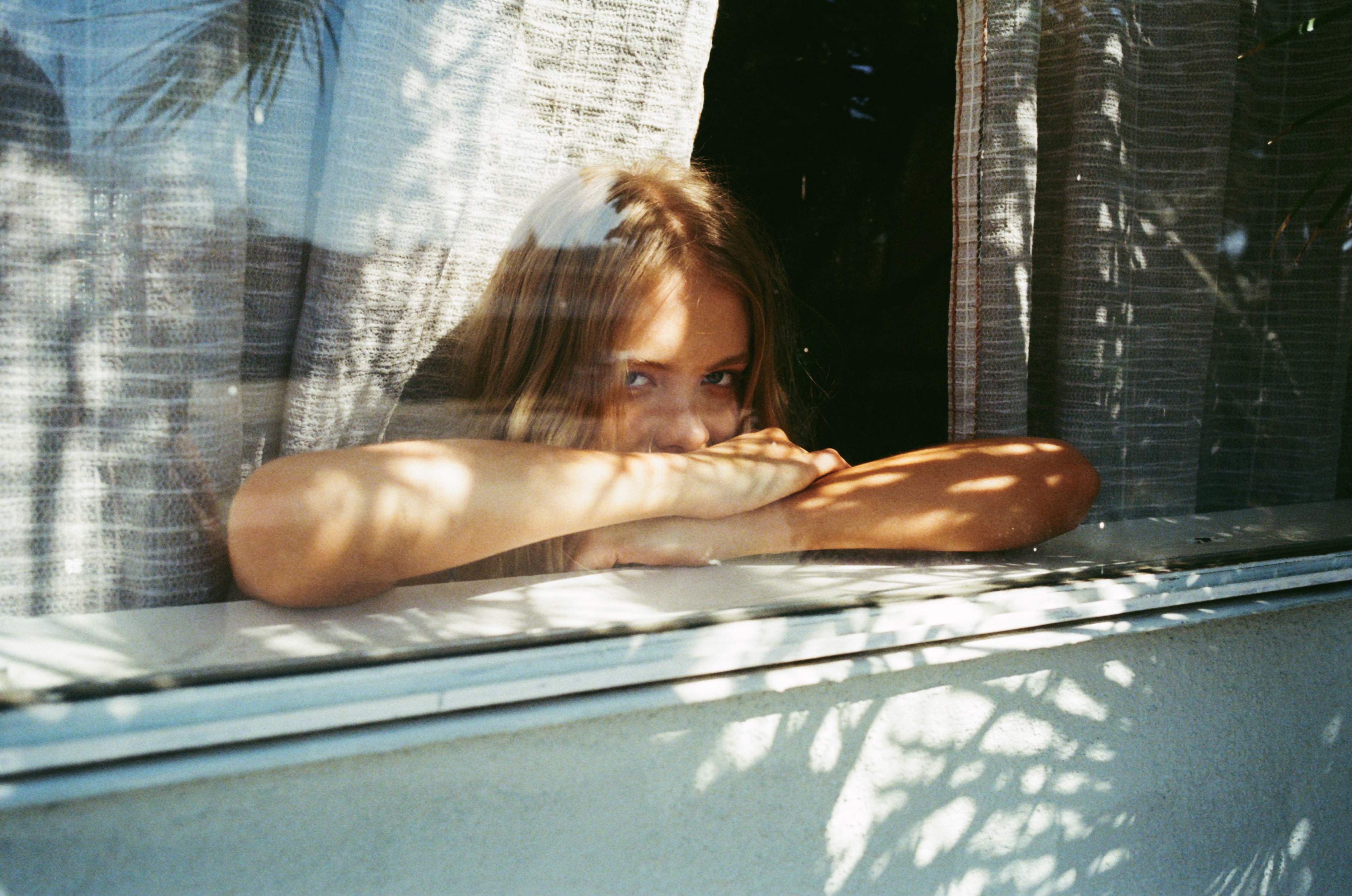 Young woman with blonde hair resting arms on a windowsill behind textured curtains, photographed by Manu Fauque.