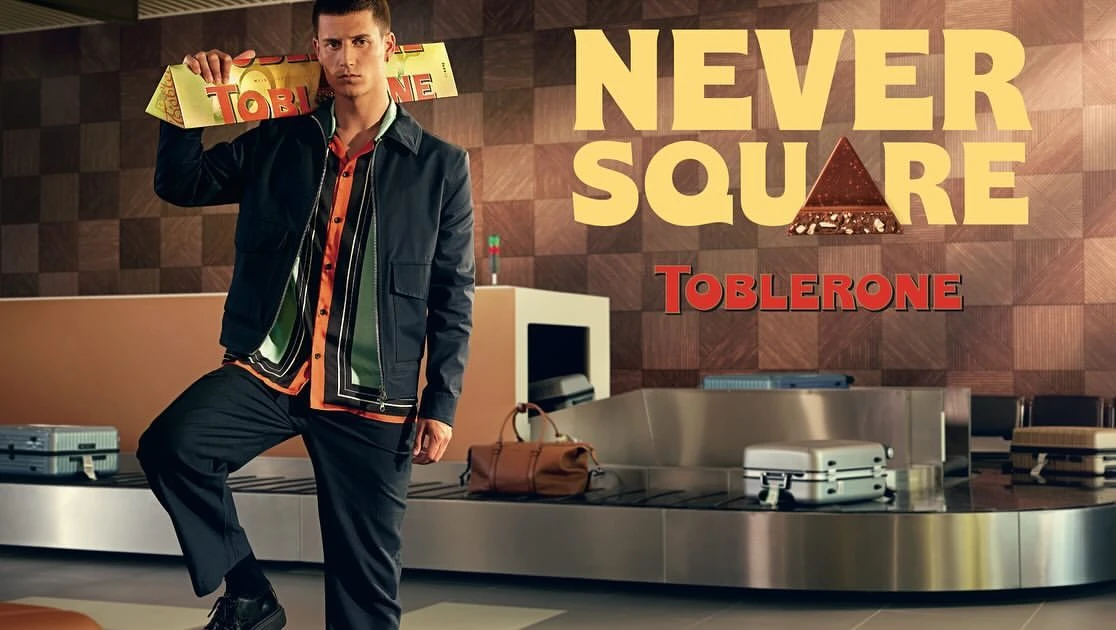 Male model poses with an oversized Toblerone bar at an airport baggage claim, advertising campaign, with luggage carousel background.