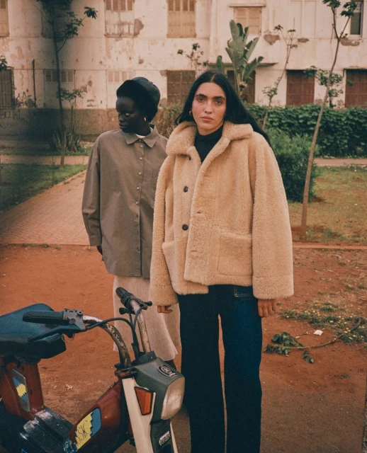 Two women in a city square pose beside a red moped; one wears a beige faux fur coat, the other a neutral overshirt and light pants.