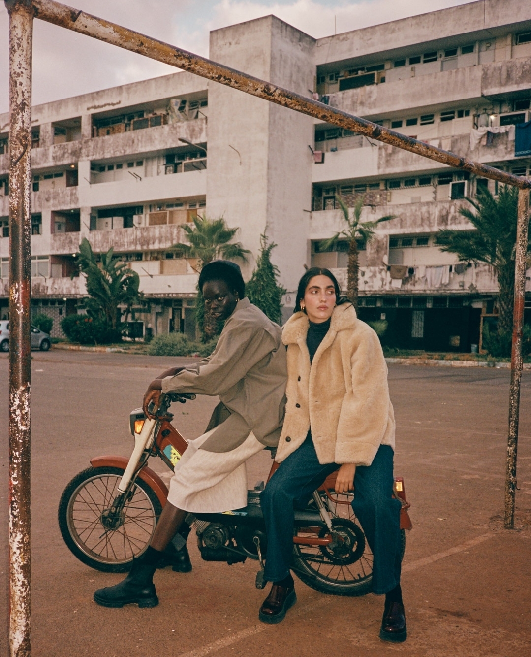 Two models pose with a red moped in front of a weathered building with palm trees, shot during daytime under overcast lighting.