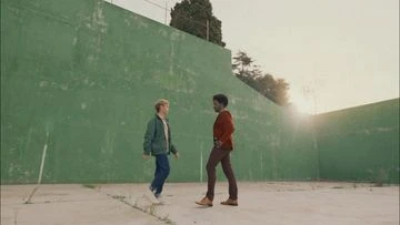 Two young men in casual clothes standing opposite each other on an outdoor court with a high green wall, under bright overcast lighting.