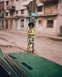 Woman in yellow patterned top and skirt posing by a wooden pole on an empty street with weathered buildings and a green car hood in the foreground.