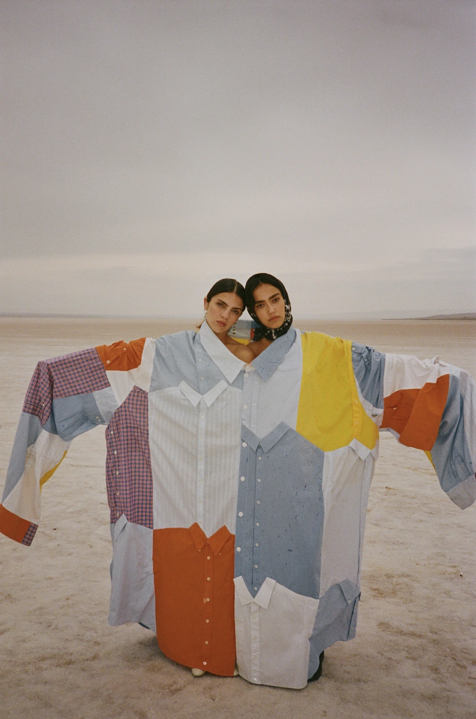 Two women wearing a giant oversized patchwork shirt made of various button-down fabrics standing on a barren landscape.