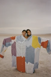 Two women wearing a giant oversized patchwork shirt made of various button-down fabrics standing on a barren landscape.