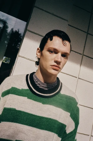 Close-up portrait of a young man with wet slicked hair wearing a green and white striped knitted sweater against tiled wall