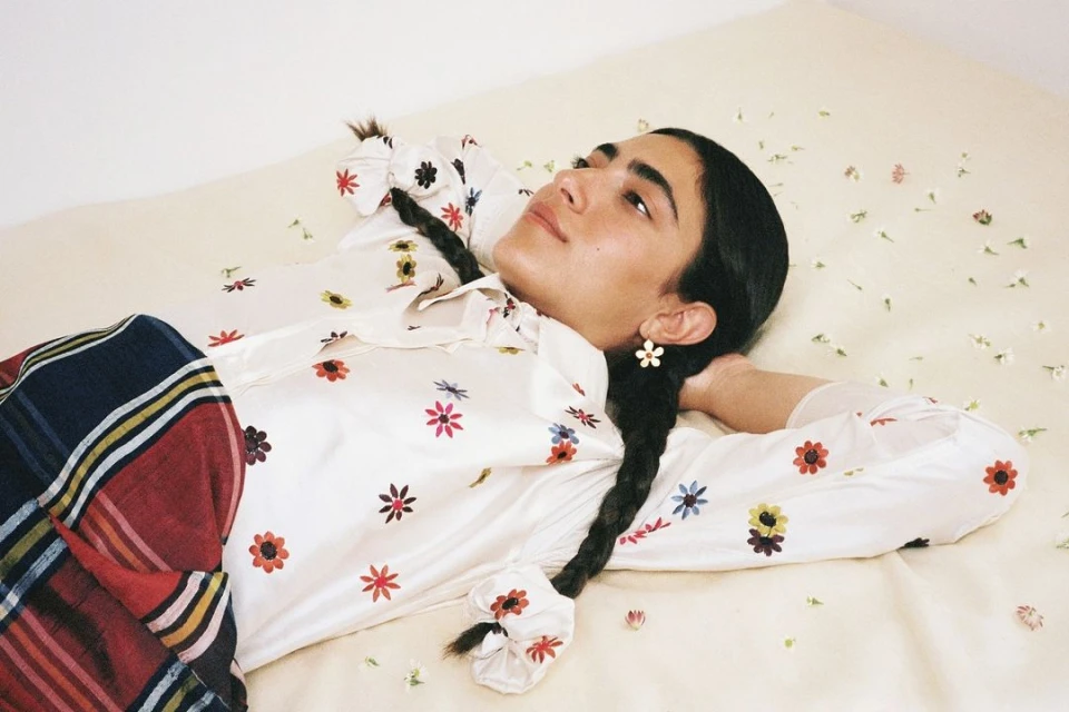 Young woman with dark hair and flower earrings relaxes on a bed with scattered flower petals, wearing a floral blouse and plaid blanket.