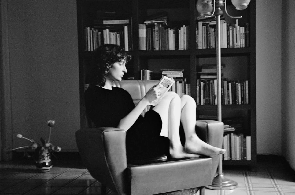 Woman in a black dress sits in a chair and reads a book; bookcase filled with books, indoor setting, black and white photo.
