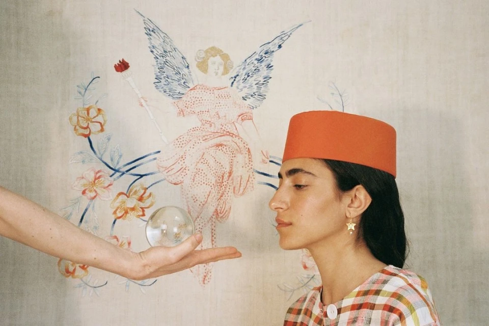 A young woman in a fez poses with a clear orb, next to an arm extending into the frame, set against a backdrop of an angel drawing.