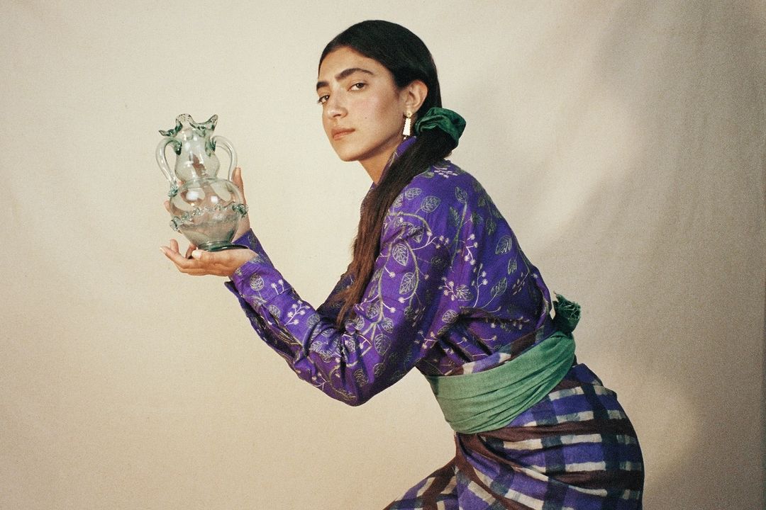 Woman in purple floral shirt holds decorative glass pitcher in a studio setting with neutral background and soft, diffused lighting.