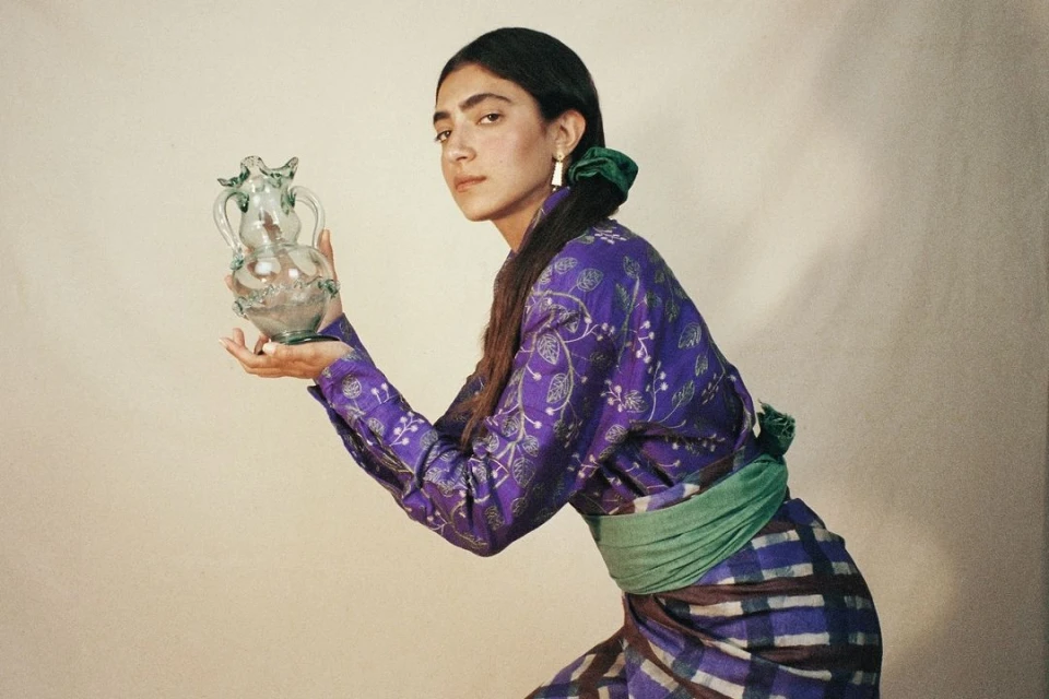 Woman in purple floral shirt holds decorative glass pitcher in a studio setting with neutral background and soft, diffused lighting.