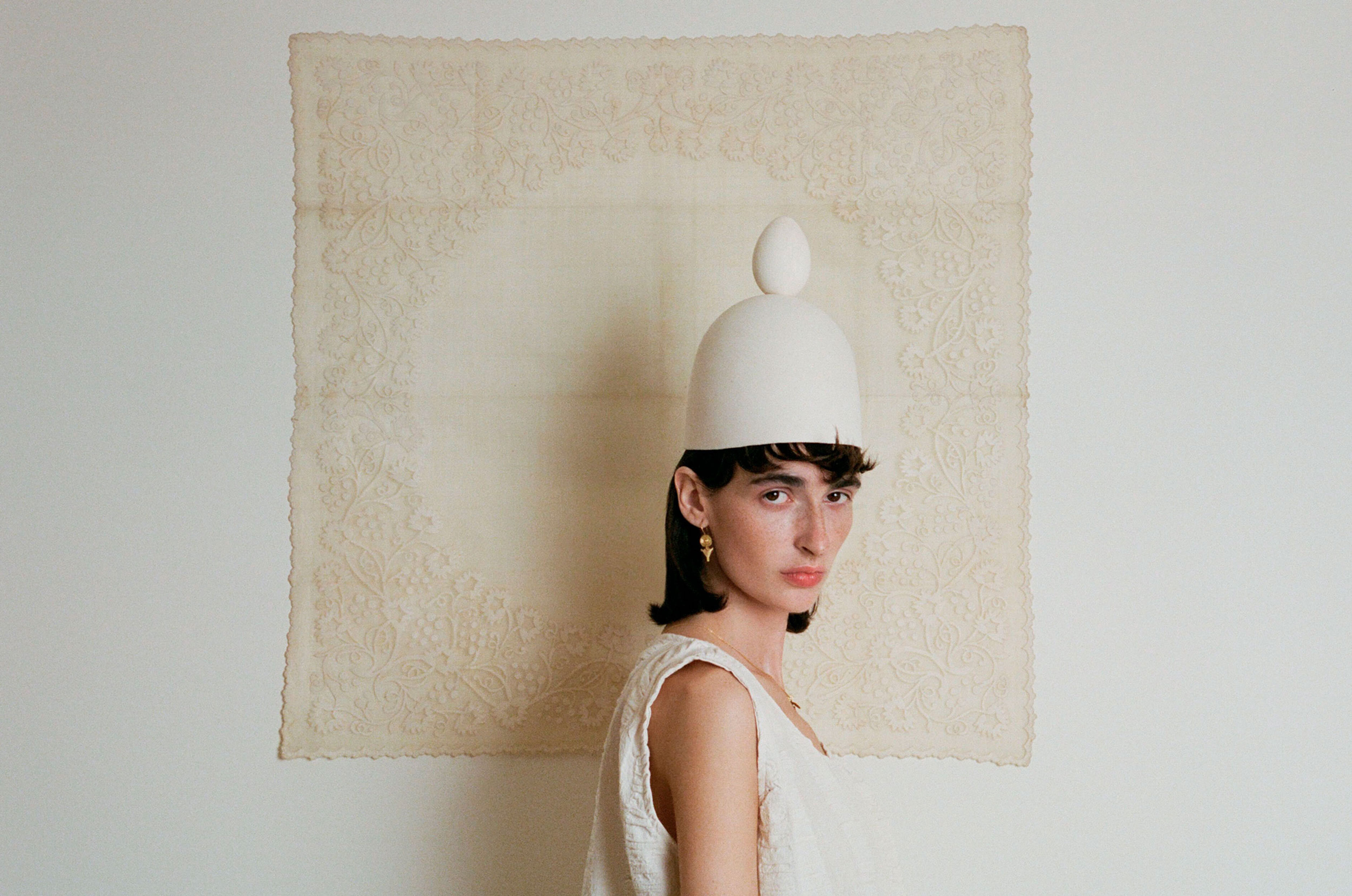 An adult female poses in front of an off-white textile backdrop while wearing a hat with egg, white dress, and jewelry in a studio setting.