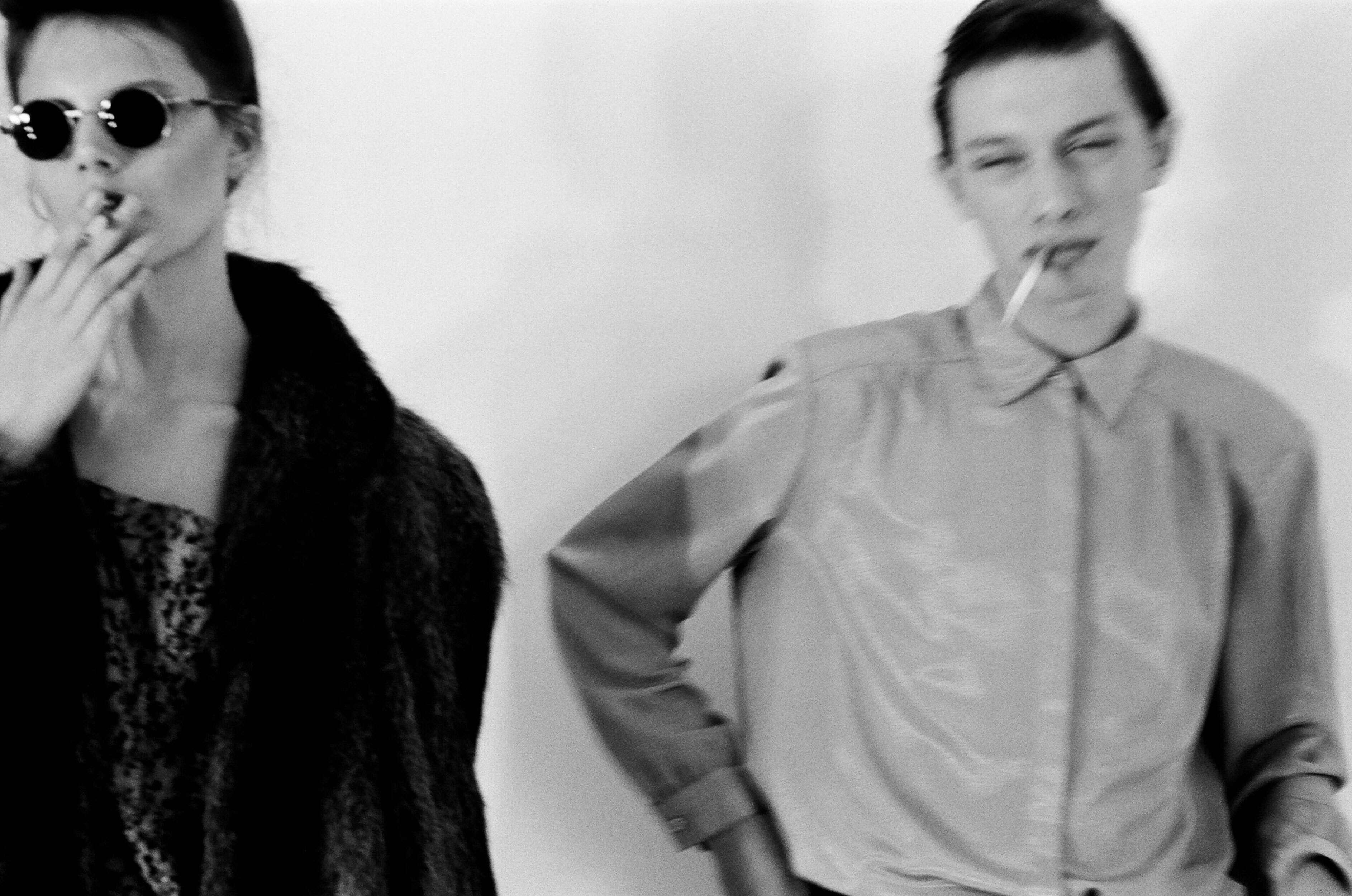 Two models in a black and white photograph with cigarettes, one wearing dark round sunglasses and a dark coat against a plain background.