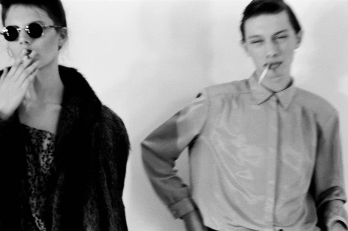 Two models in a black and white photograph with cigarettes, one wearing dark round sunglasses and a dark coat against a plain background.