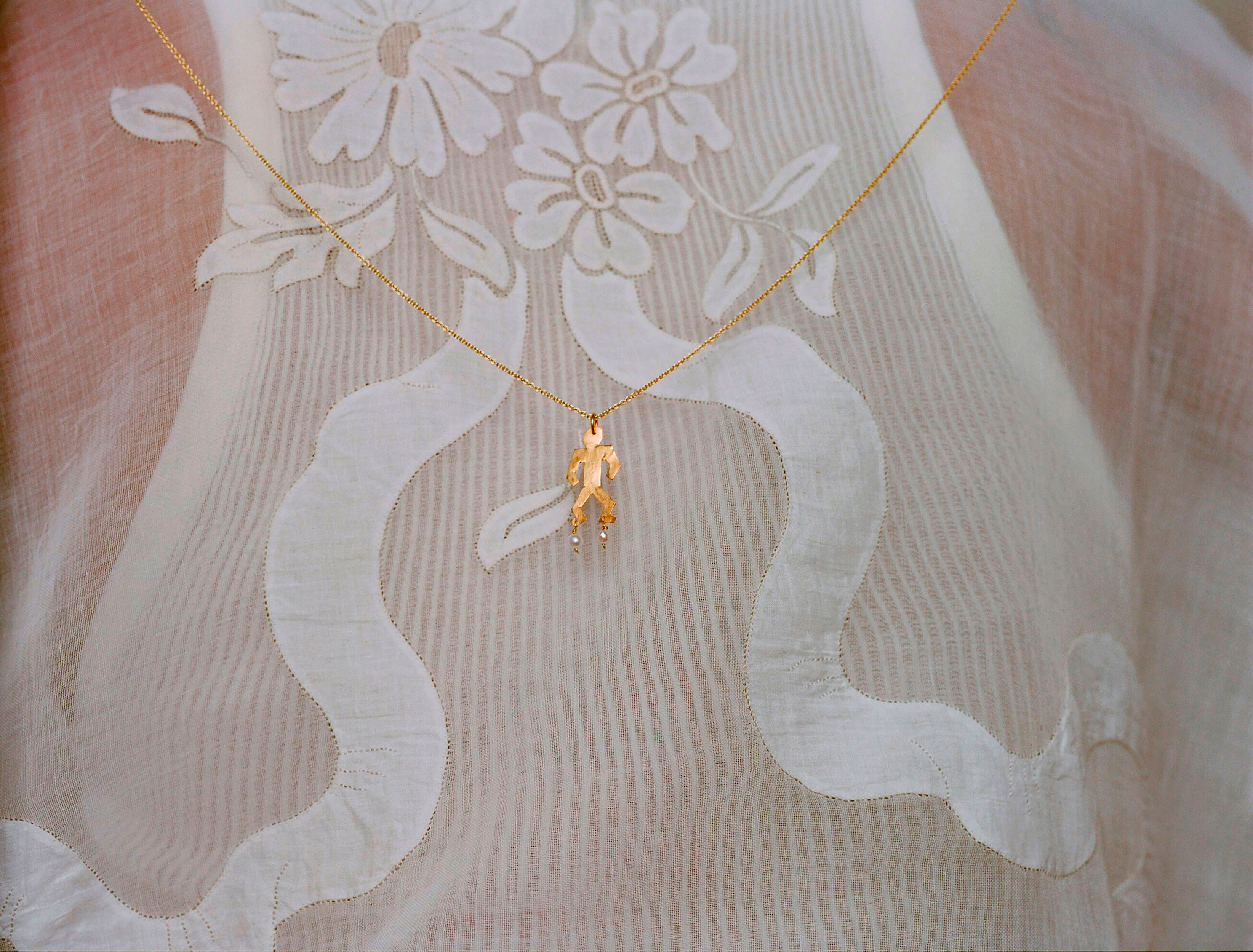 Gold necklace with a human-shaped pendant on a white, floral-patterned fabric. Delicate gold chain and small pearl-like accents.