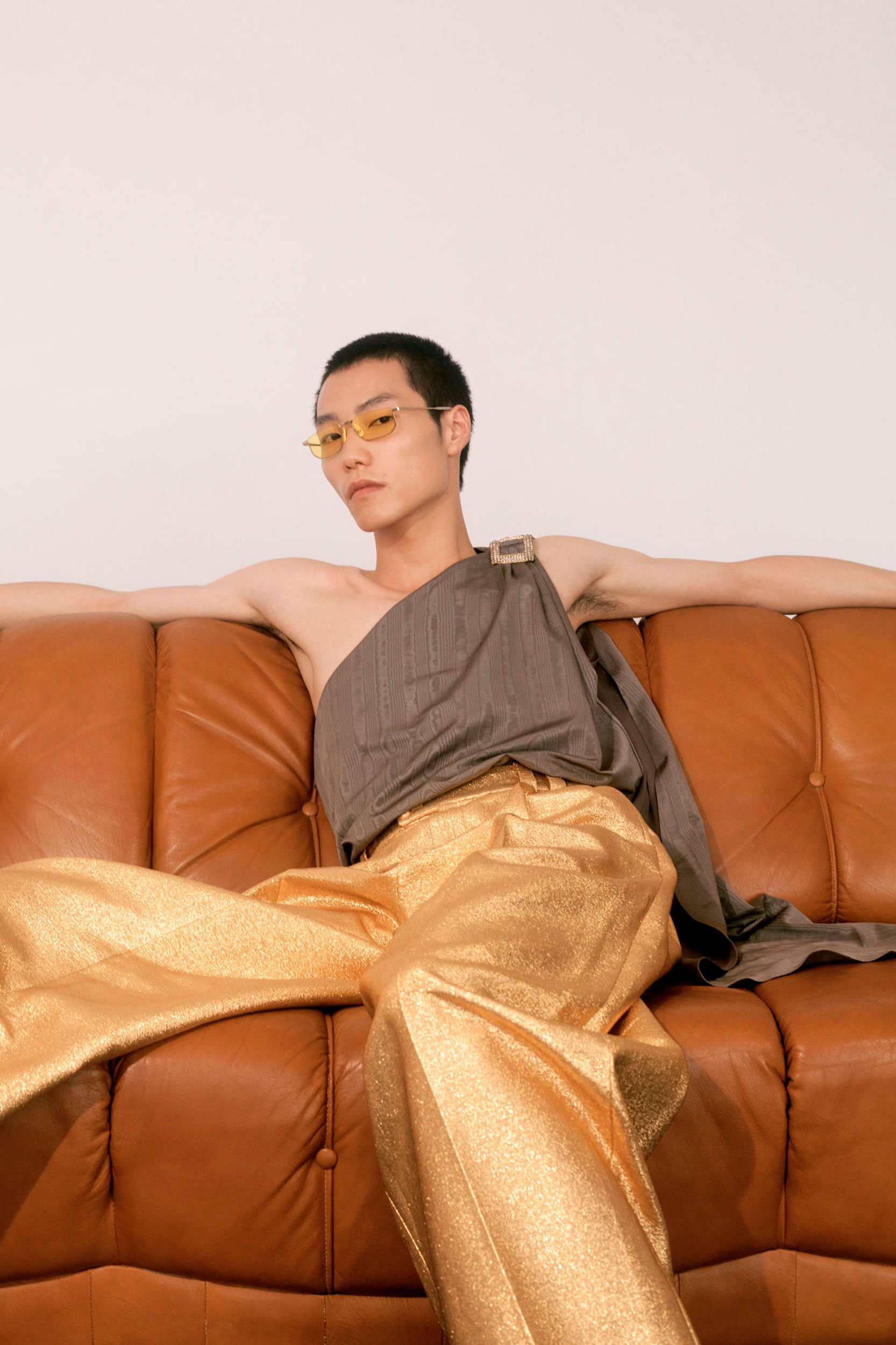 Man sitting on brown leather couch wearing gold wide-legged pants and draped top in a fashion pose in a minimalist setting.