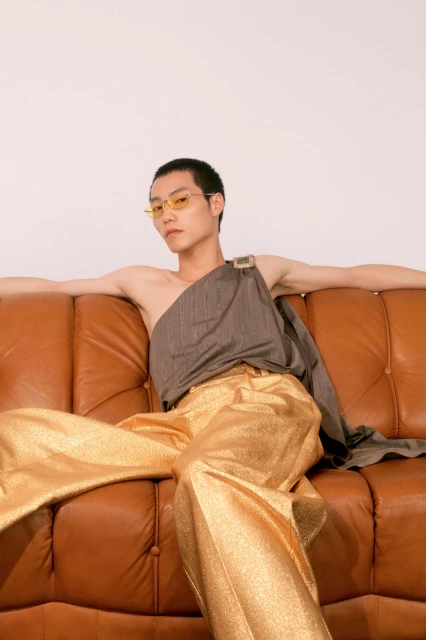 Man sitting on brown leather couch wearing gold wide-legged pants and draped top in a fashion pose in a minimalist setting.
